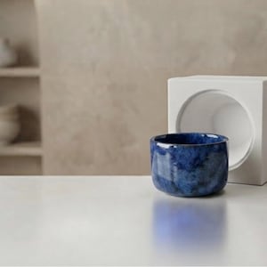 Op de afbeelding: Een kleine, ronde, diepblauwe keramische kom met een gevlekte, glanzende afwerking staat voor een wit, kubusvormig object met een grote, ronde uitsparing aan de voorzijde. De kom heeft een gladde, gebogen rand en een effen donkerblauwe basis.