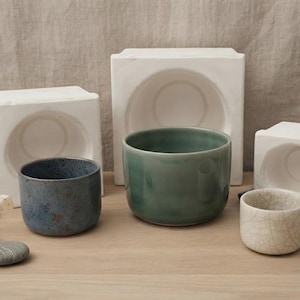 Pode incluir: Três chávenas de cerâmica em azul, verde e creme com vidrado craquelado, posicionadas à frente de três moldes de gesso branco. A chávena azul tem um acabamento salpicado, a verde é lisa e a creme apresenta um padrão de fissuras finas.