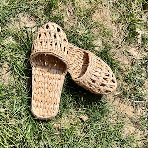 Pair of handmade woven straw sandals with open-weave uppers and flat natural soles, placed on outdoor grass. Lightweight, breathable, eco-friendly design perfect for summer beach wear or casual outdoor activities.
