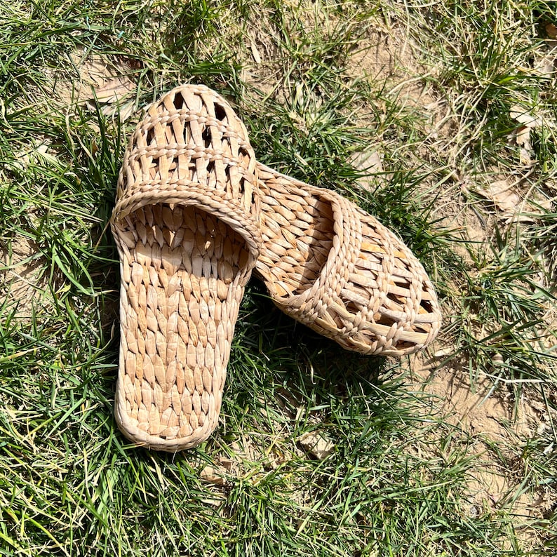 Pair of handmade woven straw sandals with open-weave uppers and flat natural soles, placed on outdoor grass. Lightweight, breathable, eco-friendly design perfect for summer beach wear or casual outdoor activities.