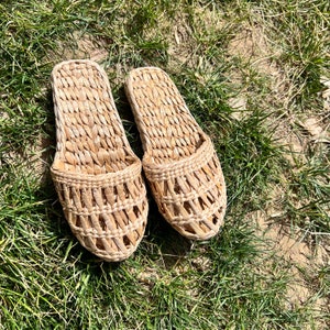 Pair of woven straw slippers with open-weave uppers and flat braided soles, placed on green grass outdoors. Lightweight, breathable, eco-friendly design ideal for summer beach wear or casual outdoor comfort.