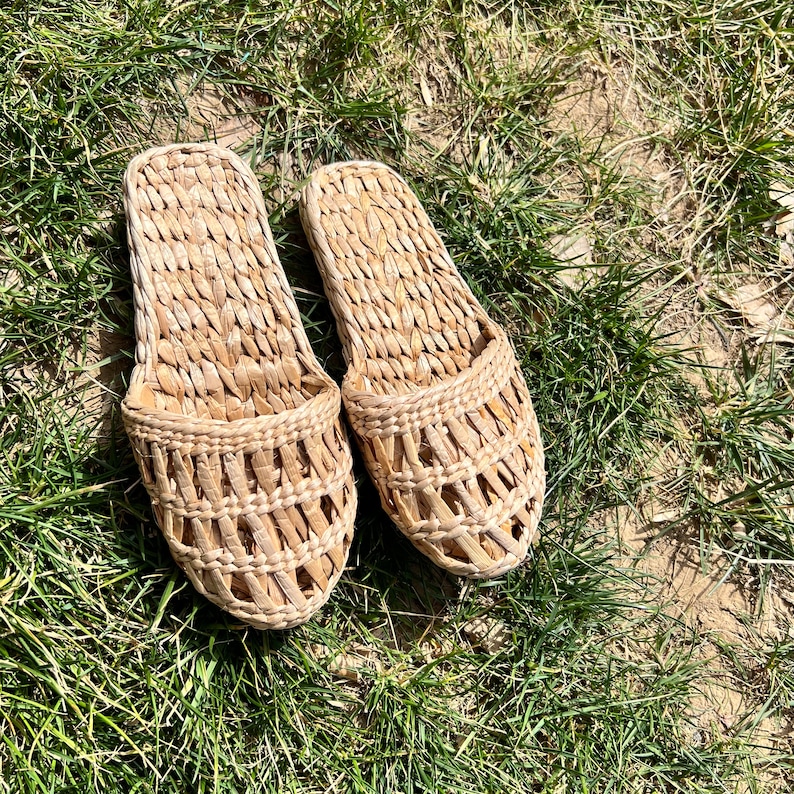 Pair of woven straw slippers with open-weave uppers and flat braided soles, placed on green grass outdoors. Lightweight, breathable, eco-friendly design ideal for summer beach wear or casual outdoor comfort.