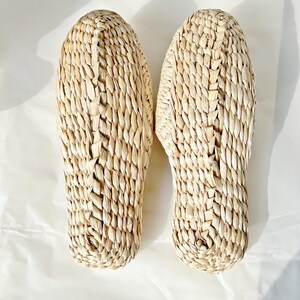 May include: A pair of natural woven straw slippers. The slippers are beige and have a woven pattern.