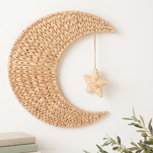May include: A woven crescent moon wall hanging with a hanging star. The moon and star are made of natural-coloured woven material. The moon is approximately 51 cm wide. The star is suspended from the moon by a thin, natural-coloured string.