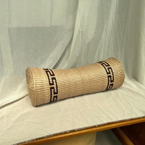 Puede incluir: Una almohada cilíndrica de color beige con un patrón geométrico en blanco y negro alrededor de los bordes. La almohada está hecha de un material tejido y está sobre una superficie blanca.