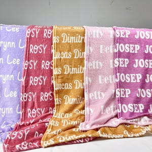 May include: Four fleece blankets with different colours and names printed on them. The blankets are folded and stacked on top of each other. The colours of the blankets are purple, red, orange, and pink.