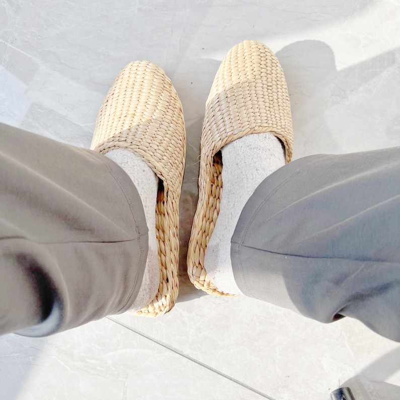 May include: A pair of woven straw slippers with a light brown color. The slippers are worn on a person's feet with white socks.