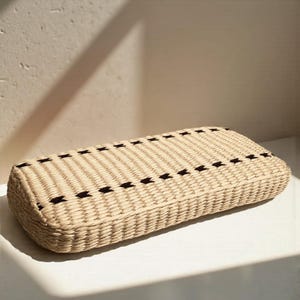 A woven basket with a textured beige pattern and black diagonal lines sits on a white surface against a plain white wall.