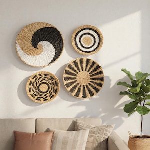 Op de afbeelding: Vier decoratieve geweven wandmanden in natuurlijke, zwarte en witte tinten. De manden hebben verschillende geometrische patronen, waaronder spiralen en concentrische cirkels. De opstelling wordt getoond op een witte muur boven een bank.