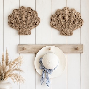 May include: Two woven seagrass wall decorations shaped like seashells mounted above a light wood coat rack with three pegs. A white straw hat with a blue and white floral patterned ribbon is hanging from the centre peg of the wooden rack.