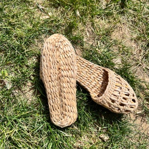 Pair of woven straw slippers with open-weave uppers and flat braided soles, one turned to show the bottom, placed on outdoor grass. Lightweight, breathable, eco-friendly design ideal for summer beach wear or casual outdoor comfort.
