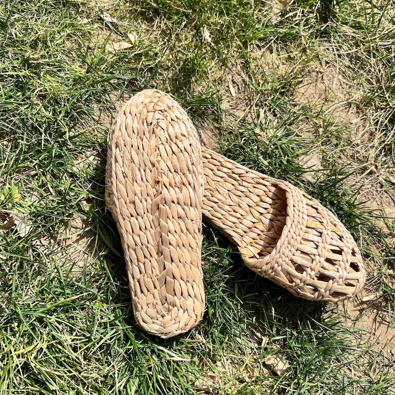 Pair of woven straw slippers with open-weave uppers and flat braided soles, one turned to show the bottom, placed on outdoor grass. Lightweight, breathable, eco-friendly design ideal for summer beach wear or casual outdoor comfort.