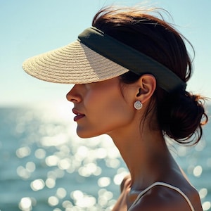 May include: A woman wearing a straw visor with a green band and a small diamond earring. The visor has a wide brim and is angled down over her face. She is looking off to the side and has her hair pulled back in a bun.