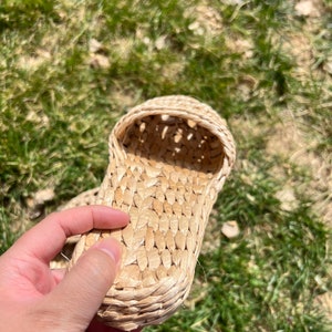 Close-up of a hand holding a woven straw slipper with open-weave upper and braided sole, shown outdoors on green grass. Lightweight, breathable, eco-friendly design ideal for summer beach wear or casual comfort.