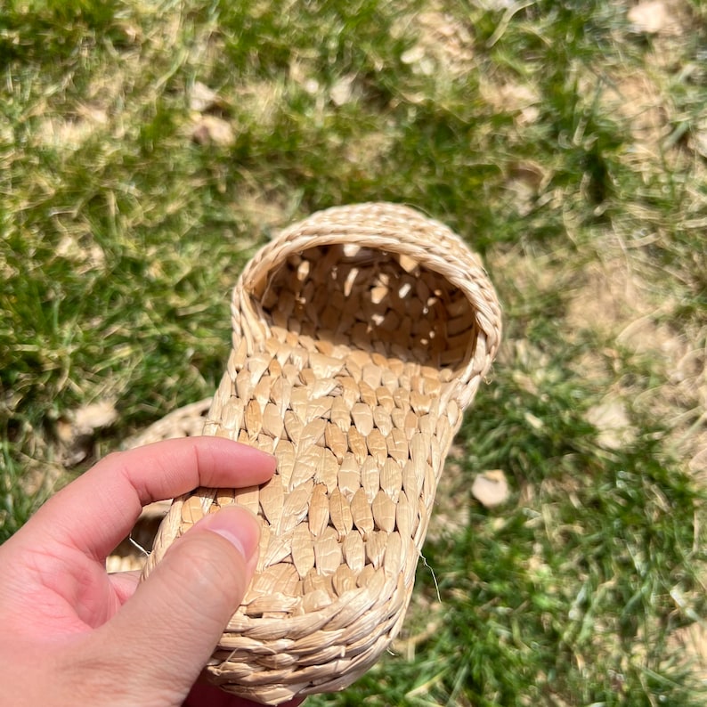 Close-up of a hand holding a woven straw slipper with open-weave upper and braided sole, shown outdoors on green grass. Lightweight, breathable, eco-friendly design ideal for summer beach wear or casual comfort.