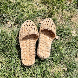 Pair of handmade woven straw slippers with open-weave uppers and flat braided soles, placed on outdoor grass. Lightweight, breathable, eco-friendly footwear ideal for summer beach wear or casual outdoor activities.