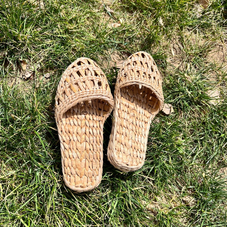 Pair of handmade woven straw slippers with open-weave uppers and flat braided soles, placed on outdoor grass. Lightweight, breathable, eco-friendly footwear ideal for summer beach wear or casual outdoor activities.