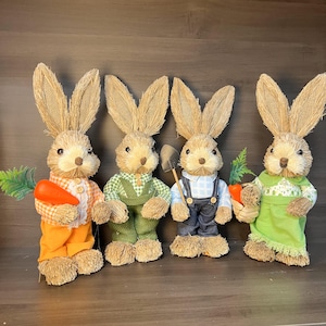 May include: Four straw bunny figurines, each wearing different clothing. One bunny holds a carrot, another holds a shovel, and another holds a basket. The bunnies are all standing on a wooden surface.