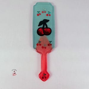May include: A decorative paddle-shaped item with a red handle and a light blue top. The top features a large cherry design with red cherries and black stems. Small cherry clusters are also scattered across the top.