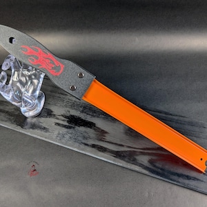 May include: A gray and orange toy car track tool. The gray handle features a red flame design and is attached to an orange track. The tool has a hole at the top of the handle and is held by a clear plastic hand.