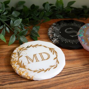 Puede incluir: Cuatro posavasos redondos con detalles de pan de oro. Los posavasos están hechos de resina y tienen diferentes diseños. Un posavasos tiene un fondo negro con una S dorada en el centro. Otro posavasos tiene un fondo rosa con un diseño floral dorado. El tercer posavasos tiene un fondo blanco con una corona floral dorada y las iniciales MD en el centro. El cuarto posavasos tiene un fondo blanco con un diseño floral dorado.