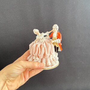 May include: A porcelain figurine depicting a couple in period attire dancing. The woman wears a pink dress with lace trim, while the man is in a red coat. The base is white with gold trim.