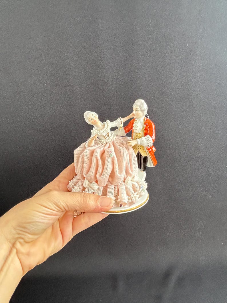 May include: A porcelain figurine depicting a couple in period attire dancing. The woman wears a pink dress with lace trim, while the man is in a red coat. The base is white with gold trim.