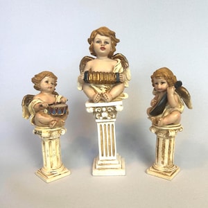May include: Three cherub figurines on decorative columns. Each cherub has golden wings and plays a musical instrument: a drum, an accordion, and a violin. The columns are white with gold detailing.