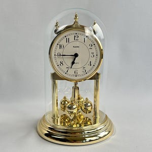 Può includere: Un orologio al quarzo color oro con quadrante bianco, numeri neri e lancette nere, racchiuso in una cupola di vetro trasparente. L'orologio ha un pendolo con sfere dorate ed è posto su una base dorata. Il quadrante dell'orologio indica "Kundo".