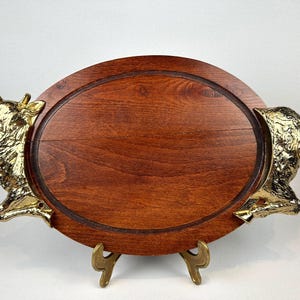 Può includere: Vassoio ovale in legno con una macchia scura, con manici a forma di testa di mucca in ottone. Il vassoio è sostenuto da un supporto in ottone. Un pezzo decorativo per servire cibo o esporre oggetti.