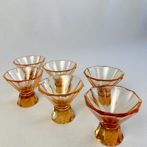 Könnte beinhalten: Ein Set aus sechs bernsteinfarbenen Glasschalen, die jeweils ein facettiertes, geometrisches Design mit einer breiten, offenen Oberseite und einem stabilen, mehrseitigen Fuß aufweisen. Das Glas verläuft von einem tiefen Orange zu einem hellen Ton.