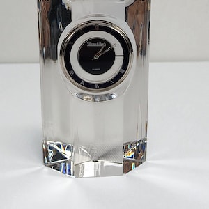 May include: A clear crystal quartz clock with a black clock face and silver hands. The clock face has Roman numerals and the brand name "Villeroy & Boch". The clock is rectangular with a faceted design.