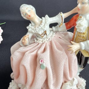 May include: A porcelain figurine depicting a couple dancing. The woman wears a pink dress with lace trim and a white blouse. The man is in a red coat and yellow vest. The figurine has a detailed design and is painted with soft colors.