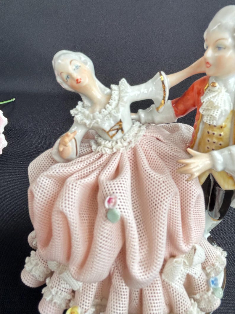 May include: A porcelain figurine depicting a couple dancing. The woman wears a pink dress with lace trim and a white blouse. The man is in a red coat and yellow vest. The figurine has a detailed design and is painted with soft colors.