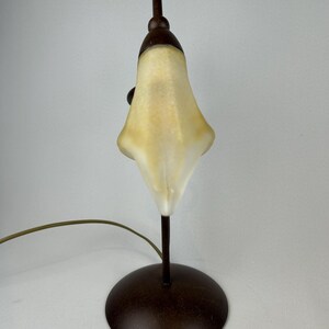 May include: A small, decorative table lamp with a bell-shaped, off-white shade. The lamp has a dark brown, circular base and a slender, matching stem. The shade is lit from within, and the lamp has a curved, brown power cord.