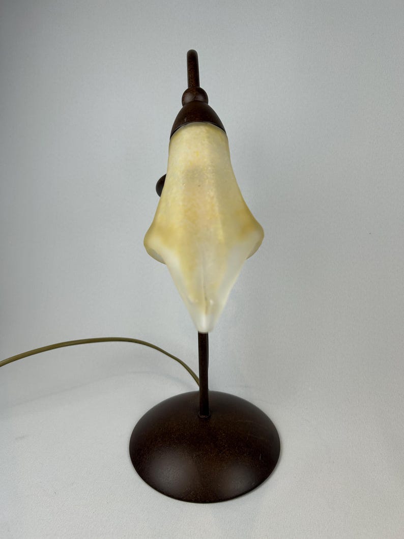 May include: A small, decorative table lamp with a bell-shaped, off-white shade. The lamp has a dark brown, circular base and a slender, matching stem. The shade is lit from within, and the lamp has a curved, brown power cord.
