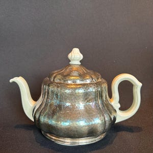 Könnte beinhalten: Eine silberfarbene Teekanne mit weißem Griff, Ausguss und Deckelknauf. Die Teekanne hat einen abgerundeten Körper mit geriffeltem Design. Der Deckel hat einen dekorativen Knopf. Die Teekanne steht auf einem silberfarbenen Sockel.