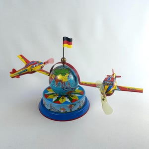 Puede incluir: Un juguete de hojalata de estilo vintage con dos aviones rojos y amarillos unidos a un globo terráqueo central. El globo está coronado por una pequeña bandera alemana. La base es circular, decorada con un patrón de estrella y ferias.