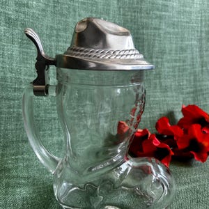 Op de afbeelding: Een bierpul van helder glas in de vorm van een laars met een metalen deksel dat eruitziet als een cowboyhoed. Het deksel heeft een touwachtige textuur rond de rand en een metalen duimhendel bevestigd aan het glazen handvat aan de zijkant.