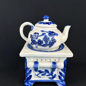 Könnte beinhalten: Eine weiße Keramik-Teekanne mit blauen Blumenmustern, die auf einem passenden herdähnlichen Sockel steht. Die Teekanne hat einen gebogenen Griff und Ausguss, und der Sockel weist dekorative Blättermuster und simulierte Herd-Elemente auf. Das Design ist im Vintage-Stil gehalten.