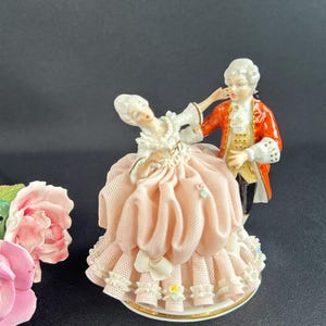 May include: A porcelain figurine depicting a couple in 18th-century attire. The woman wears a pink dress with lace trim, while the man is in a red coat. The base is white with a gold trim. A pink rose is in the background.