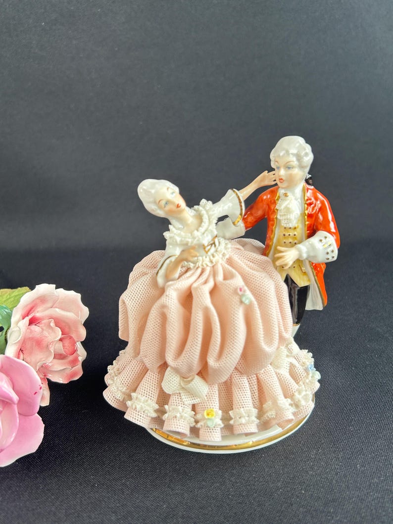 May include: A porcelain figurine depicting a couple in 18th-century attire. The woman wears a pink dress with lace trim, while the man is in a red coat. The base is white with a gold trim. A pink rose is in the background.
