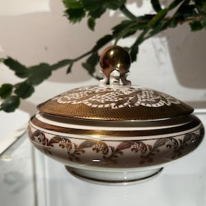 Puede incluir: Una caja de porcelana blanca para alhajas con una tapa dorada y diseños florales intrincados. La tapa tiene un pomo dorado con un diseño decorativo.