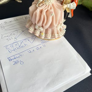 May include: A porcelain figurine of a dancing couple, the woman in a pink dress with a ruffled skirt and the man in a red coat. The base is gold-trimmed. A piece of paper with handwritten notes is in the foreground.