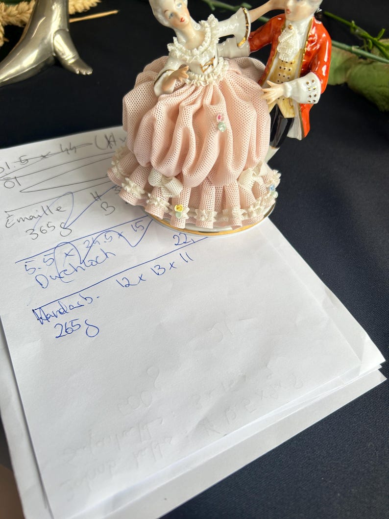 May include: A porcelain figurine of a dancing couple, the woman in a pink dress with a ruffled skirt and the man in a red coat. The base is gold-trimmed. A piece of paper with handwritten notes is in the foreground.