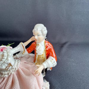 May include: A porcelain figurine depicting a couple in period attire. The man wears a red jacket with gold accents, a white ruffled shirt, and a powdered wig. The woman is in a pink dress with lace details. The figurine is a decorative item.