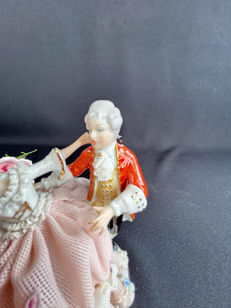 May include: A porcelain figurine depicting a couple in period attire. The man wears a red jacket with gold accents, a white ruffled shirt, and a powdered wig. The woman is in a pink dress with lace details. The figurine is a decorative item.