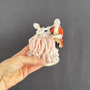May include: A porcelain figurine depicting a couple in formal attire dancing. The woman wears a pink dress with lace trim, while the man is in a red coat. The base is white with a gold trim. The figurine is a decorative item.