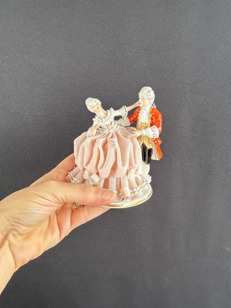 May include: A porcelain figurine depicting a couple in formal attire dancing. The woman wears a pink dress with lace trim, while the man is in a red coat. The base is white with a gold trim. The figurine is a decorative item.