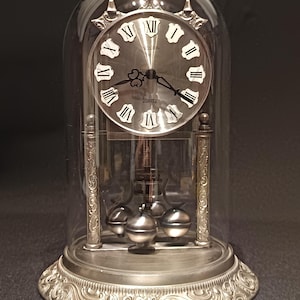 Könnte beinhalten: Eine silberfarbene Quarz-Jahresuhr unter einer Glaskuppel. Die Uhr hat ein kreisförmiges Zifferblatt mit schwarzen römischen Ziffern, verzierten Zeigern und dem Wort QUARTZ unter der Mitte. Ein rotierendes Pendel mit vier Kugeln befindet sich unter dem Zifferblatt, gestützt von zwei dekorativen Säulen auf einem runden Sockel mit geprägten Schnörkeln.